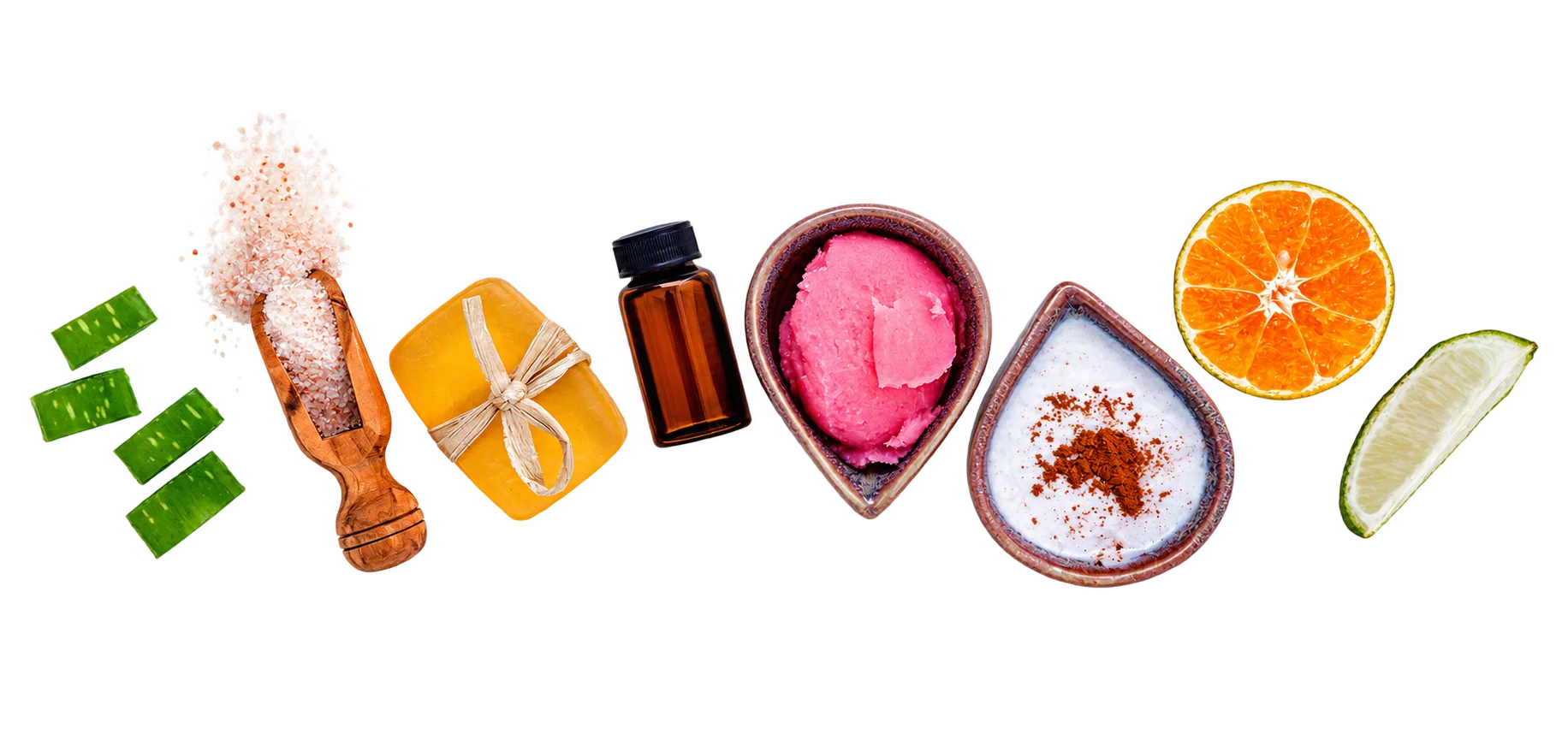 Des morceaux d’aloe vera, du sel rose dans une cuillère en bois, un savon au miel, un flacon ambré, des bols de gommage rose et de poudre, une orange et un citron vert sont alignés sur une surface blanche.