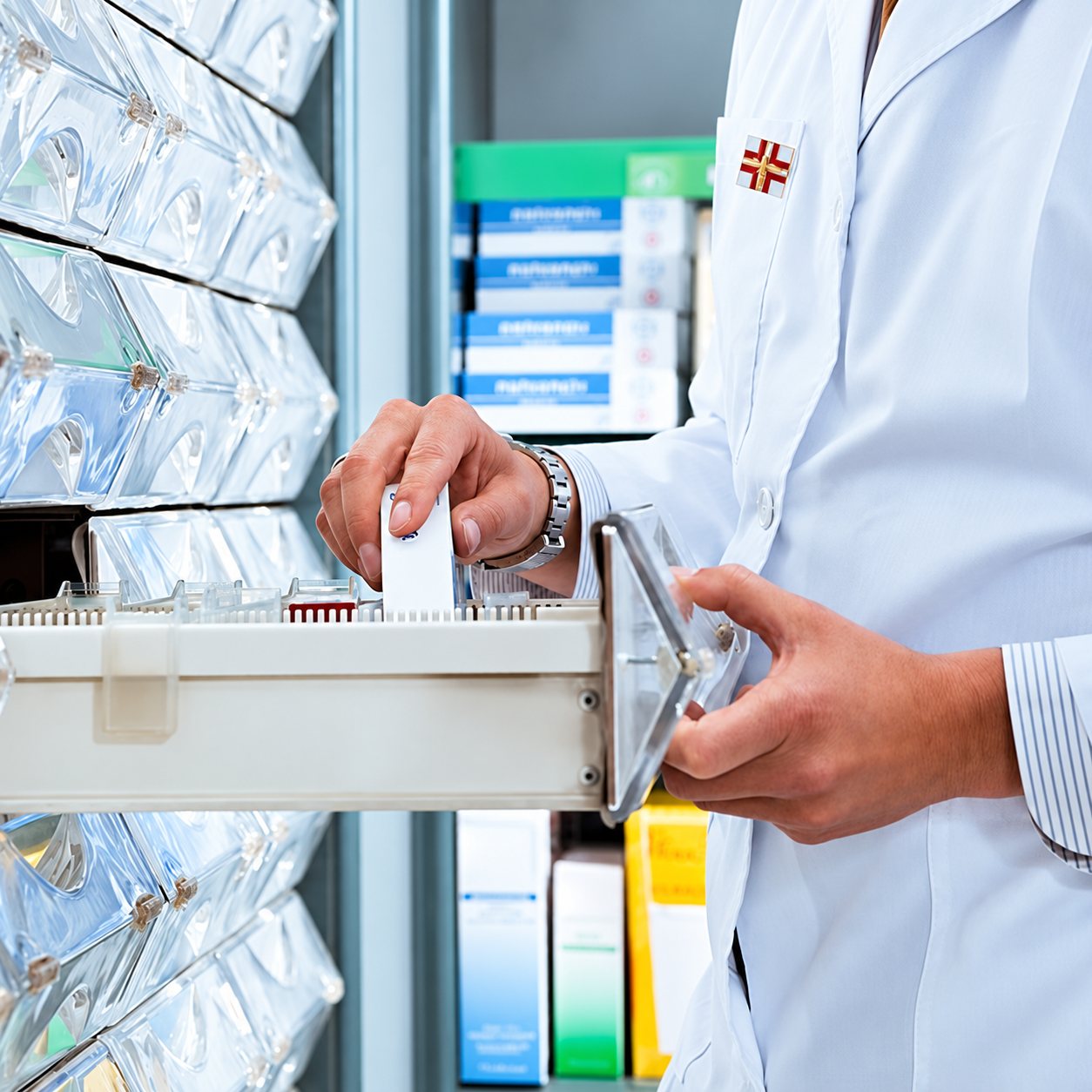 Un pharmacien en blouse blanche retire une boîte de médicaments d’un tiroir coulissant dans l’officine, avec des étagères de produits en arrière-plan.