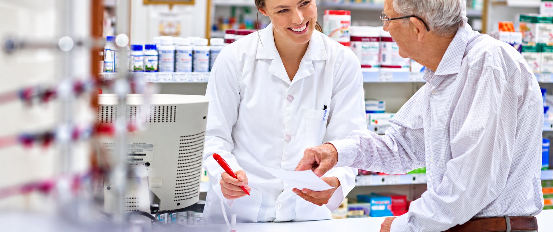 Une pharmacienne en blouse blanche sourit en montrant une ordonnance à un client âgé au comptoir d’une pharmacie.