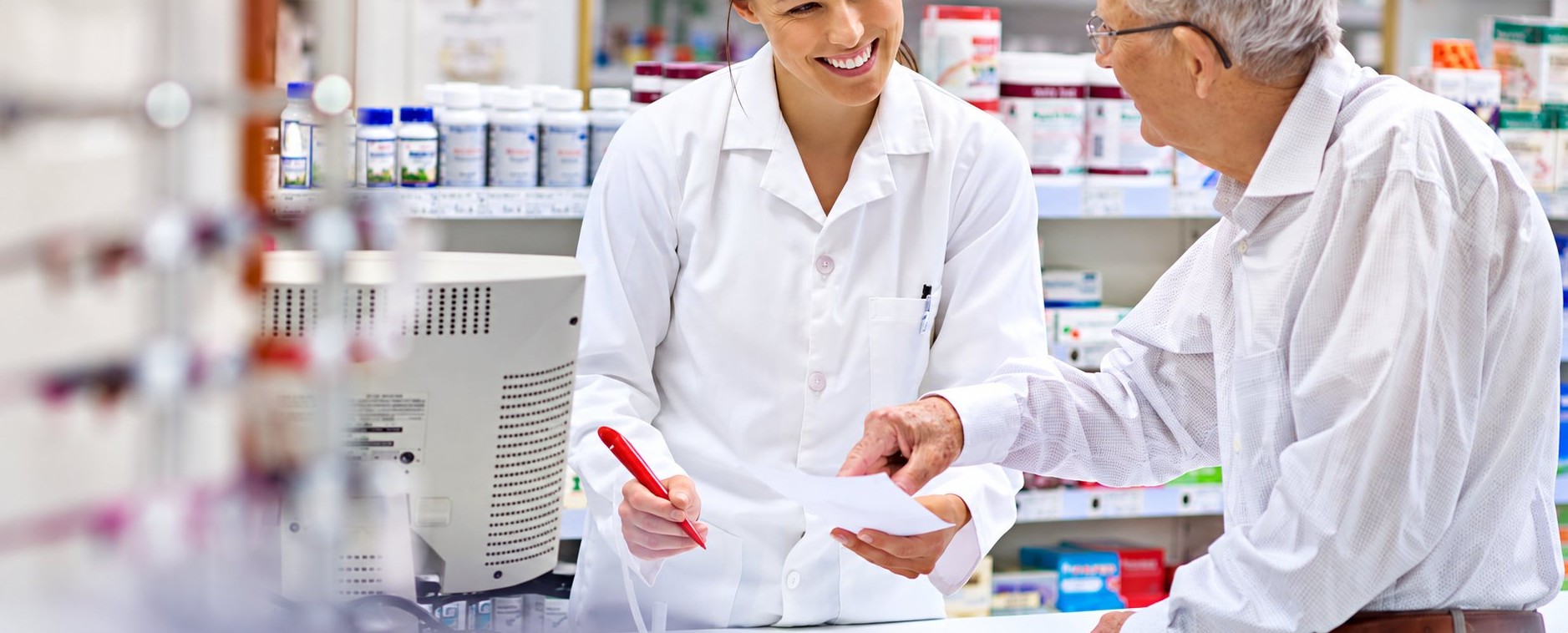 Une pharmacienne en blouse blanche sourit en tenant un stylo rouge et une ordonnance pendant qu’elle conseille un client âgé au comptoir.